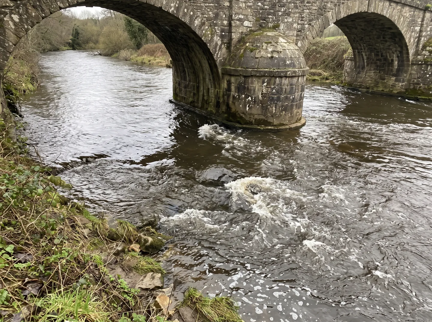 A river flowing past the stone pier of an old bridge, with swirling eddies and ripples forming downstream of the pier